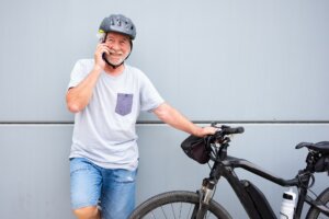 Smiling senior man with helmet using phone leaning against gray wall enjoying activity with bike